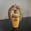 Lattafa Afeef by Lattafa 100ml EDP — Gold fluted bottle with two ornate gold peacocks adorned with colorful gems, standing on a black surface against a light grey wall.