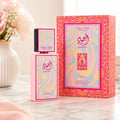 Lattafa Ajwad Pink 2 Pink Creamy Elixir — Botol minyak wangi pink dan kotak sepadan dengan corak abstrak berwarna-warni, diletakkan di atas permukaan marmar dengan pasu bunga pink di latar belakang.