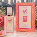 Lattafa Ajwad Pink 2 Pink Creamy Elixir — Botol minyak wangi merah jambu dan kotak corak orennya dipamerkan bersama bunga merah jambu dan latar belakang bandar yang kabur.