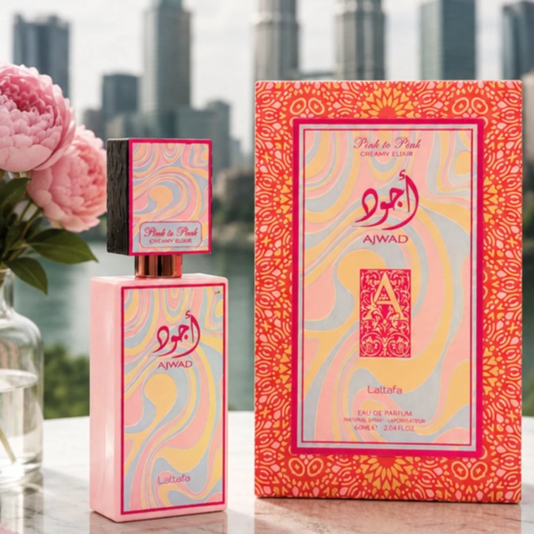 Lattafa Ajwad Pink 2 Pink Creamy Elixir — Botol minyak wangi merah jambu dan kotak sepadan dengan rekaan pastel berpusar, berlatar belakang langit bandar yang kabur dan bunga peoni merah jambu.