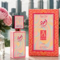 Lattafa Ajwad Pink 2 Pink Creamy Elixir — Botol minyak wangi merah jambu dan kotak sepadan dengan rekaan pastel berpusar, berlatar belakang langit bandar yang kabur dan bunga peoni merah jambu.