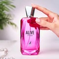Maison Alhambra Alive Now — Pink faceted glass perfume bottle held by a hand with manicured nails, with a green plant and beaded necklace in the background. | Maison Alhambra Alive Now — Botol kaca faset merah jambu dipegang oleh tangan berkuku manikur, dengan tumbuhan hijau dan kalung manik di latar belakang.