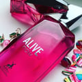 Maison Alhambra Alive Now by Maison Alhambra 100ml — A bright pink perfume bottle with silver text "ALIVE now" and a deer logo, surrounded by colorful abstract earrings and a cherry earring. | Maison Alhambra Alive Now by Maison Alhambra 100ml — Botol minyak wangi merah jambu terang dengan teks perak "ALIVE now" dan