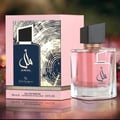 Ard Al Zaafaran Amal Ard Al Zaafaran 100ml Wanita — Kotak merah jambu corak bijian kayu dan botol biru gelap dengan kaligrafi Arab, bersebelahan botol minyak wangi kaca merah jambu bertutup hitam.