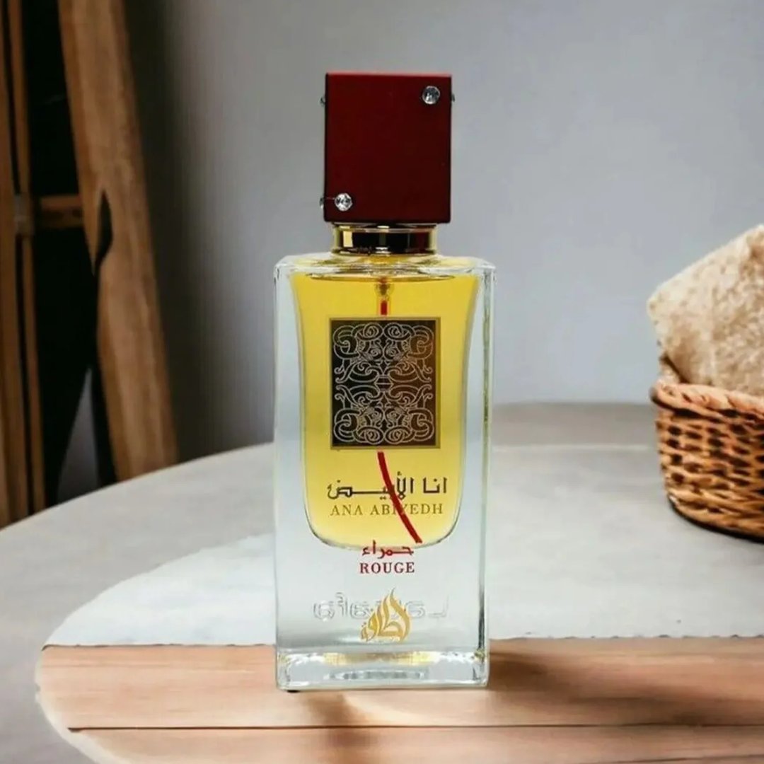 Lattafa Ana Abiyedh Rouge Lattafa 100ml EDP — Botol minyak wangi kaca jernih berisi cecair kuning, penutup merah gelap, dan kaligrafi Arab pada label, diletakkan di atas permukaan kayu dengan latar belakang bakul dan tuala yang kabur.