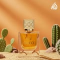 Lattafa Aura by Lattafa 60ml EDP — A clear glass perfume bottle with golden liquid and a decorative gold cap sits on sand, surrounded by prickly pear cacti and rocks against an orange background. | Lattafa Aura by Lattafa 60ml EDP — Botol minyak wangi kaca jernih dengan cecair keemasan dan penutup emas hiasan duduk di
