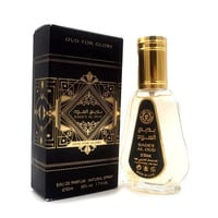 Ard Al Zaafaran Badee Al Oud Eau De Parfum 50ml — Black and gold patterned box next to a clear glass perfume bottle with a gold cap, on a white background. | Ard Al Zaafaran Badee Al Oud Eau De Parfum 50ml — Kotak hitam dan emas bermotif di sebelah botol minyak wangi kaca jernih bertutup emas, di atas latar belakang