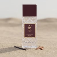 Bujairami Billion Dollar by Bujairami 100ml Extrait — Clear glass perfume bottle with a maroon cap, set on sand with a vanilla bean and sandalwood pieces.