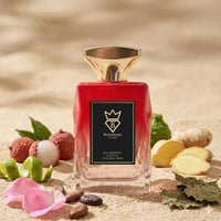 Bujairami Boujee by Bujairami 100ml EDP — Red faceted glass bottle with gold cap, surrounded by lychees, ginger slices, lime zest, rose petals, and cocoa beans on a sandy background.