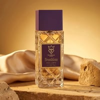 Bujairami Breathless by Bujairami 100ml Extrait — Clear faceted glass bottle with gold label, purple cap, on a wooden surface with draped silk and rocks.