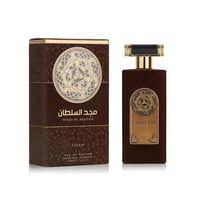 Asdaaf Majd Al Sultan by Asdaaf 100ml EDP — A brown velvet-textured perfume bottle and its matching box are displayed side-by-side against a white background. Both feature a circular gold emblem with an intricate knot design and Arabic text. | Asdaaf Majd Al Sultan by Asdaaf 100ml EDP — Sebotol dan kotak minyak wangi