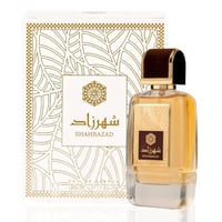 Ard Al Zaafaran Shahrazad Eau De Parfum 100ml — Botol kaca jernih berisi cecair keemasan, berpenutup coklat dan emas, diletakkan di sebelah kotak putih berhias corak daun emas.