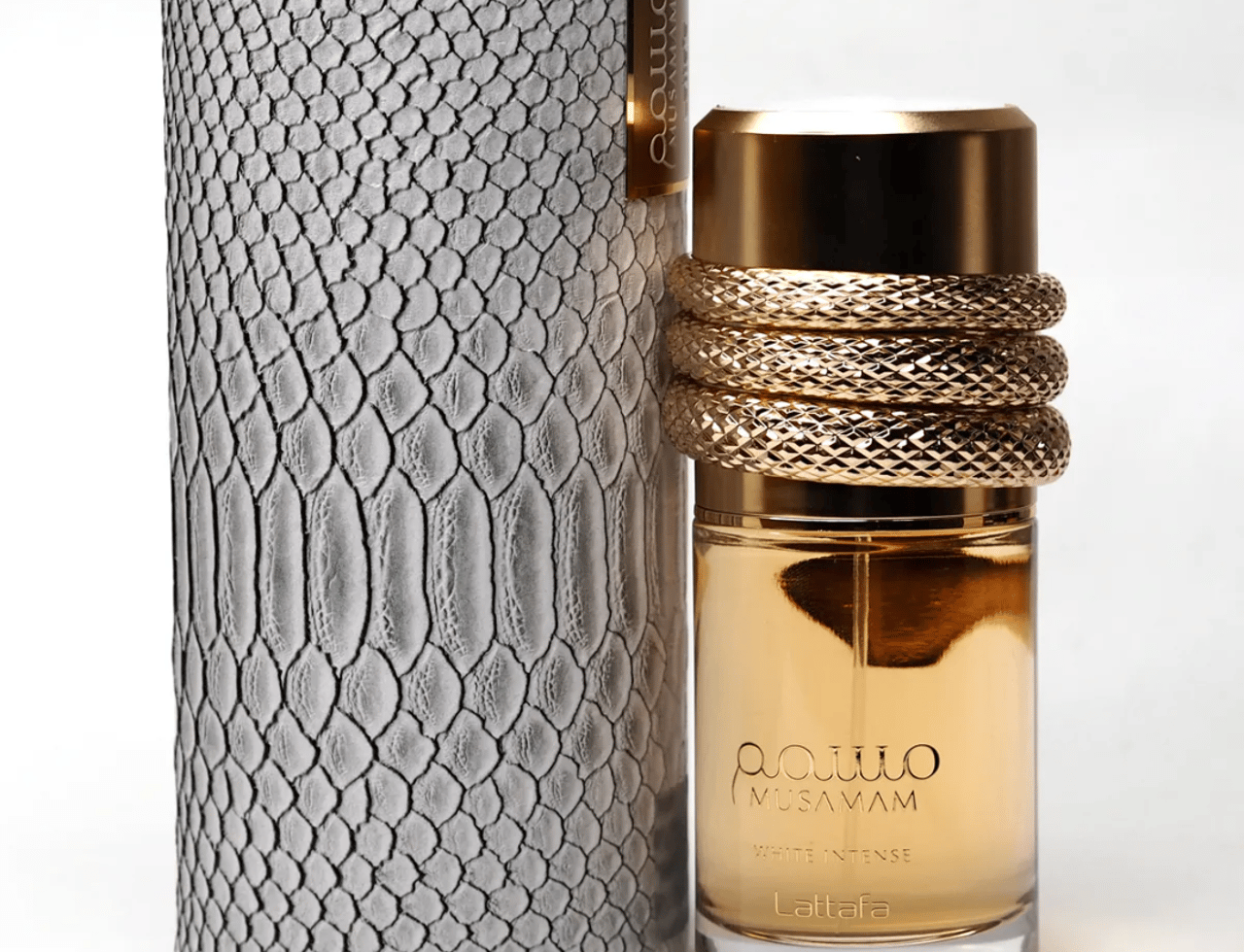 Lattafa Musamam Intense by Lattafa 100ml EDP — Kotak silinder kelabu bertekstur tinggi berdiri di sebelah botol minyak wangi kaca jernih dengan perincian emas dan reka bentuk gegelung seperti ular di sekeliling lehernya.
