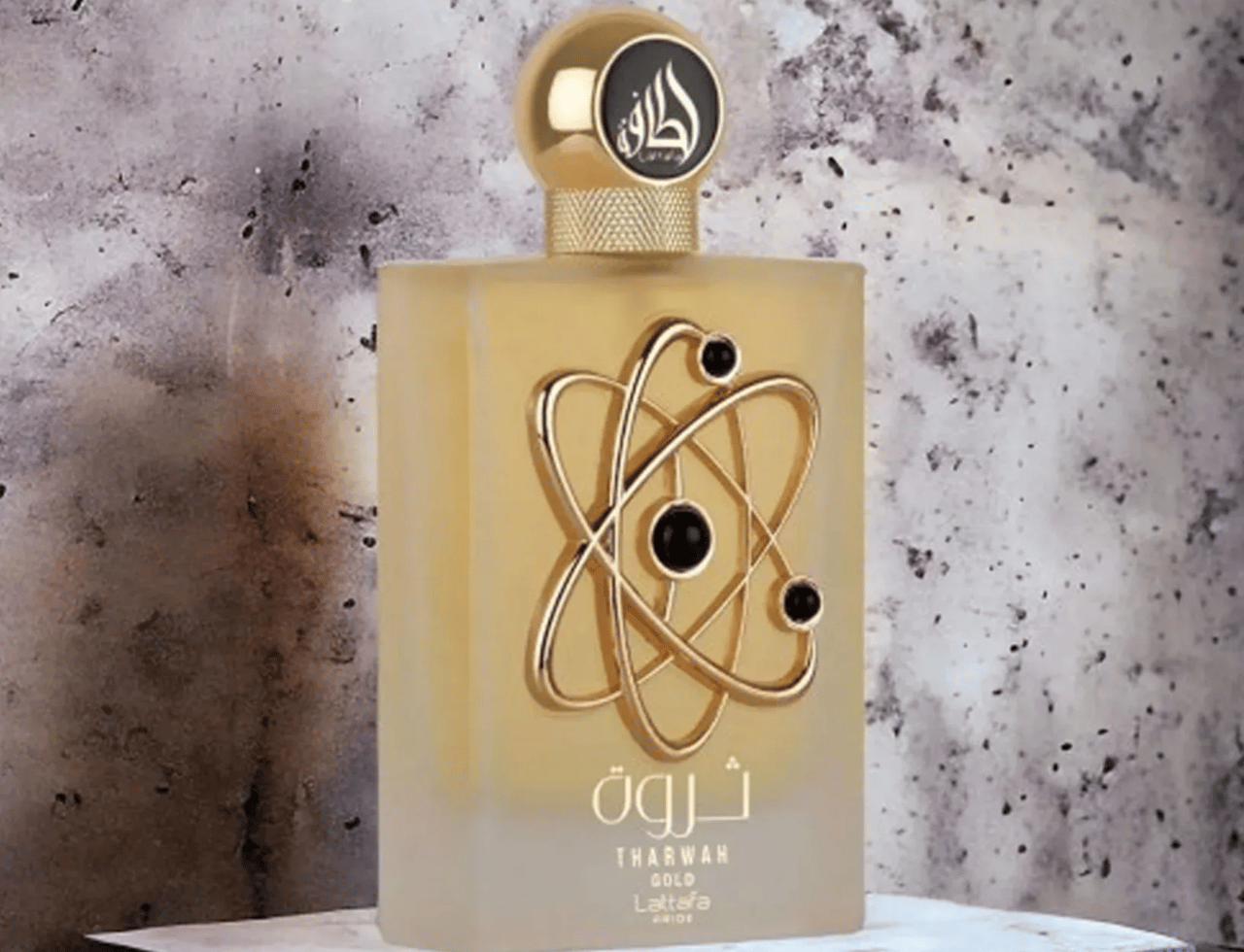 Lattafa Pride Tharwah Gold by Lattafa Pride 100ml EDP — Botol minyak wangi kaca emas beku dengan lambang seperti atom emas dan sfera hitam, berlatar belakang kelabu bertekstur.