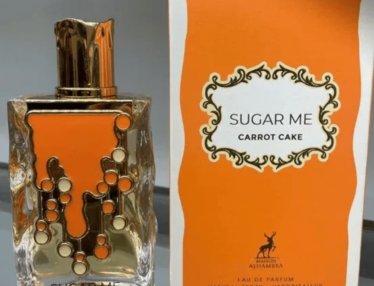 Maison Alhambra Sugar Me Carrot Cake — Clear glass perfume bottle with gold cap and orange decorative elements, next to its orange and white product box.