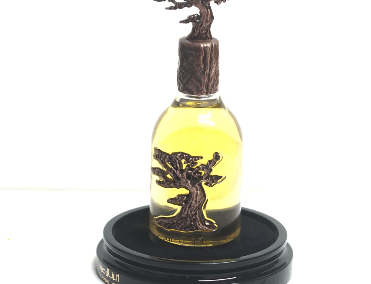 Lattafa Pride Eternal Oud by Lattafa Pride 100ml EDP — Yellow perfume in a clear glass bottle with a bronze tree-shaped cap and a bronze tree emblem on the bottle, displayed on a black circular base with text.
