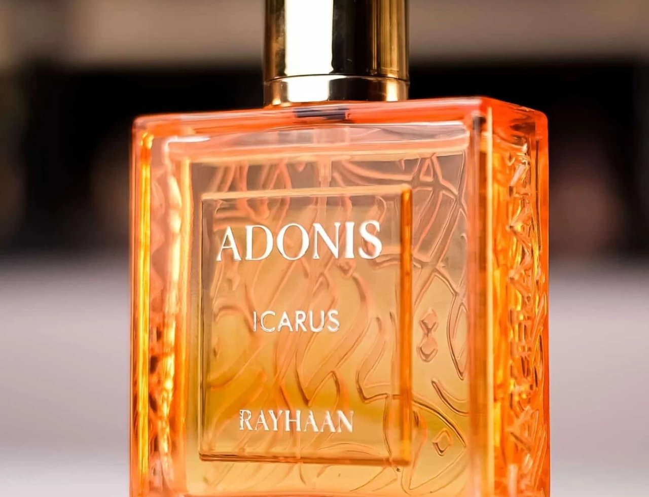Rayhaan Adonis Icarus Rayhaan Perfume 100ml — A square, amber-colored glass perfume bottle with a gold cap, featuring embossed Arabic calligraphy and text "ADONIS ICARUS RAYHAAN" on the front. | Rayhaan Adonis Icarus Rayhaan Perfume 100ml — Botol kaca berbentuk segi empat berwarna ambar dengan penutup emas