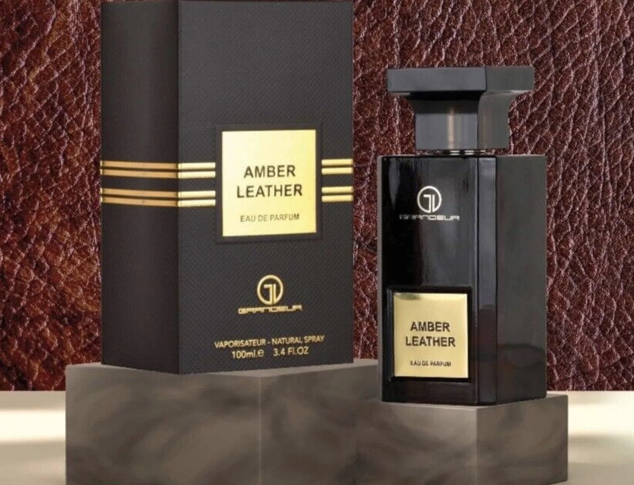 Grandeur Amber Leather 100ml by Grandeur Perfumes — A black perfume box and bottle are displayed on dark grey marble stands against a textured brown leather background. | Kotak dan botol minyak wangi hitam Grandeur Amber Leather 100ml dipamerkan di atas tapak marmar kelabu gelap berlatarkan latar belakang kulit coklat