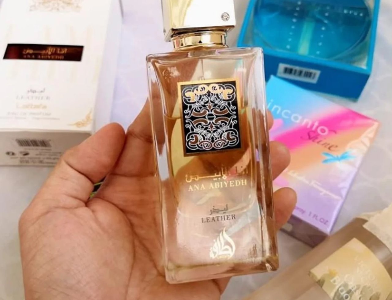 Lattafa Ana Abiyedh Leather 60ml by Lattafa Perfumes — Botol wangian kaca jernih dengan penutup emas berkilauan, dipegang di tangan. Kotak putih Lattafa dan kotak wangian Incanto Shine kelihatan di latar belakang.