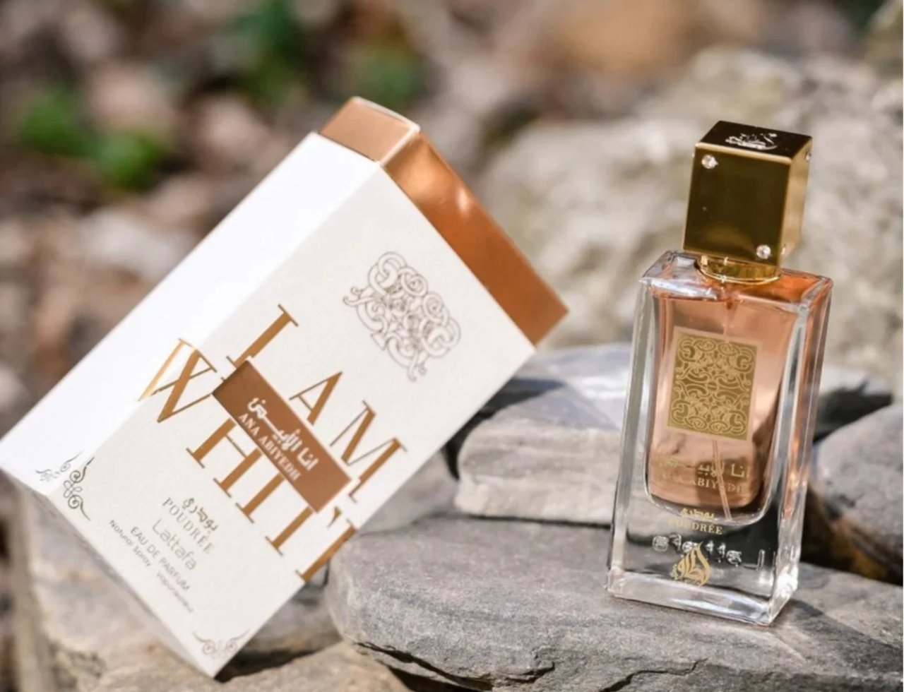Lattafa Ana Abiyedh Poudree 60ml — A white and gold perfume box tilted on its side next to a clear glass perfume bottle with a gold cap, both resting on grey rocks with a blurred natural background. | Lattafa Ana Abiyedh Poudree 60ml — Kotak minyak wangi putih dan emas diletakkan senget di sebelah botol minyak wangi