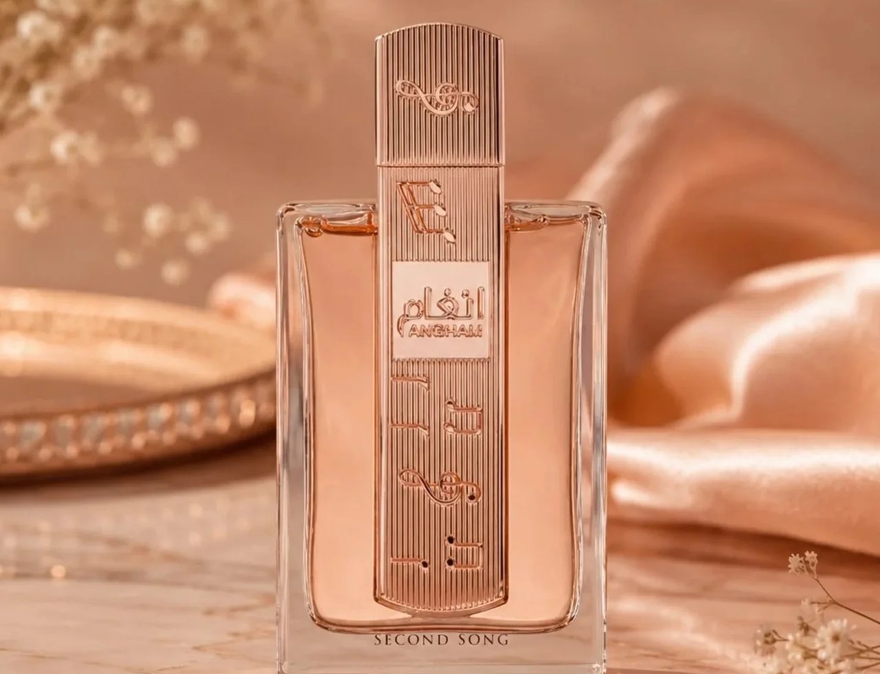 Lattafa Angham Second Song by Lattafa — Rose gold fluted cap and label on a clear glass perfume bottle, filled with peachy liquid, set against a soft peach satin background with delicate white flowers.