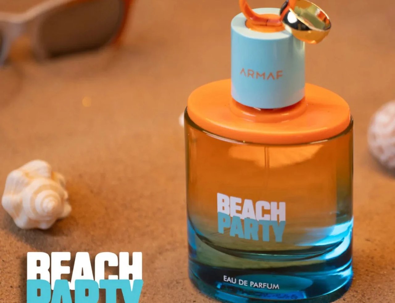 Armaf Beach Party Eau De Parfum — Botol wangian berwarna jingga dan biru di atas pasir, dengan cengkerang dan cermin mata hitam di latar belakang