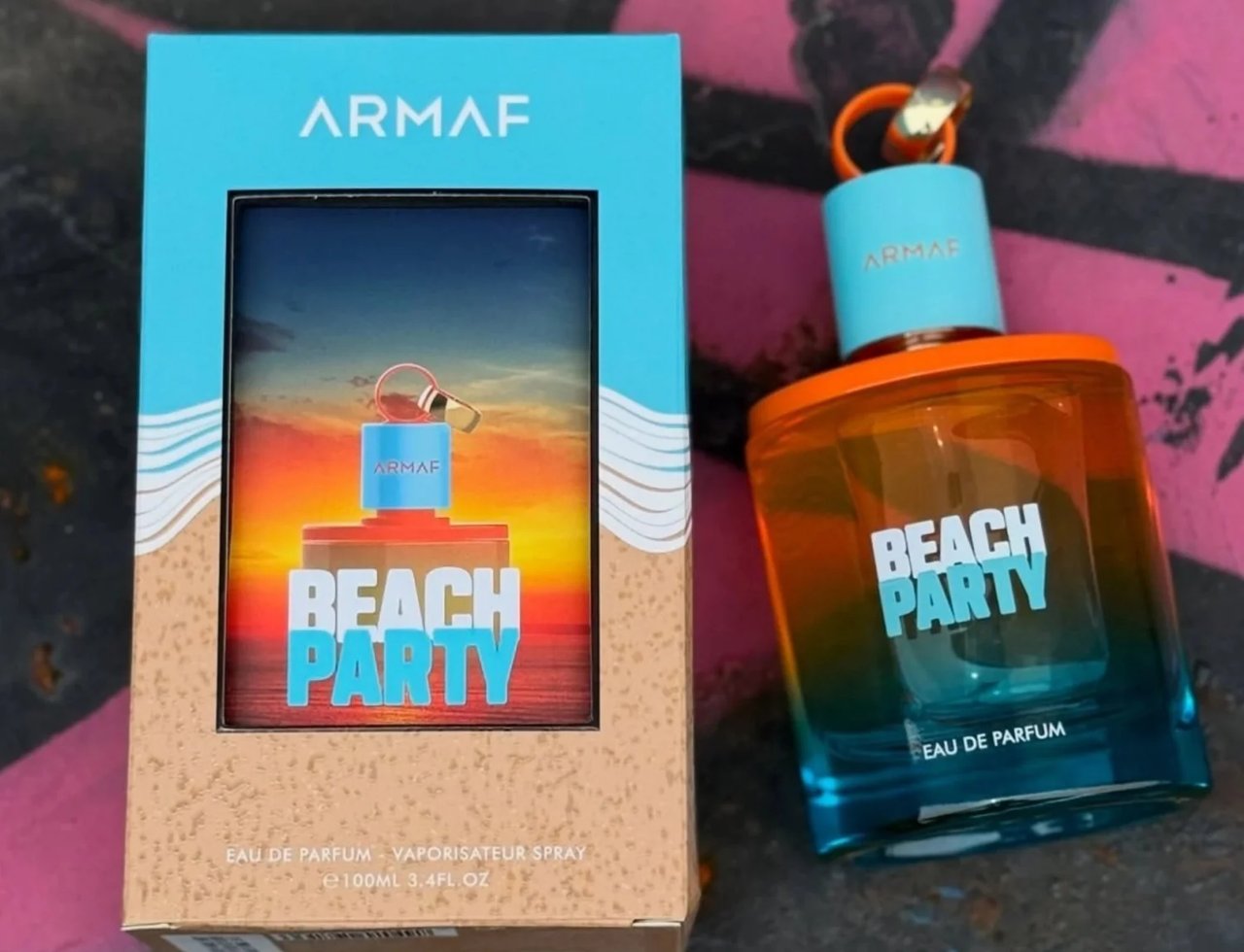 Armaf Beach Party Eau De Parfum 100ml — A blue and tan box with a beach sunset scene on the front is placed next to a gradient orange and blue glass perfume bottle with a light blue cap and orange ring | Armaf Beach Party Eau De Parfum 100ml — Kotak biru dan coklat dengan pemandangan matahari terbenam pantai di