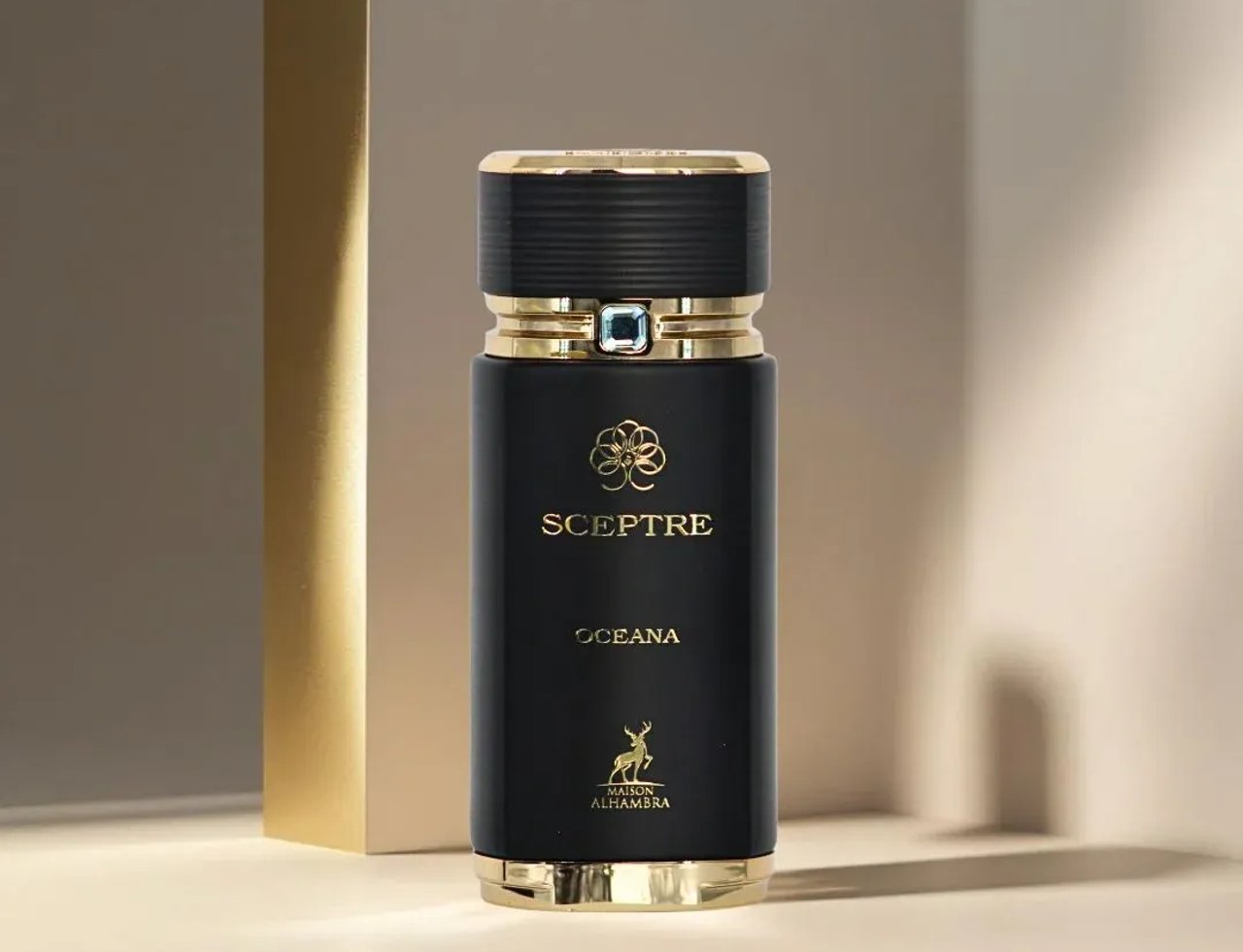 Maison Alhambra Sceptre Oceana — Botol minyak wangi hitam dengan perincian emas dan permata biru pada penutup, berdiri di sebelah tiang emas berlatar belakang lembut dan berbayang.