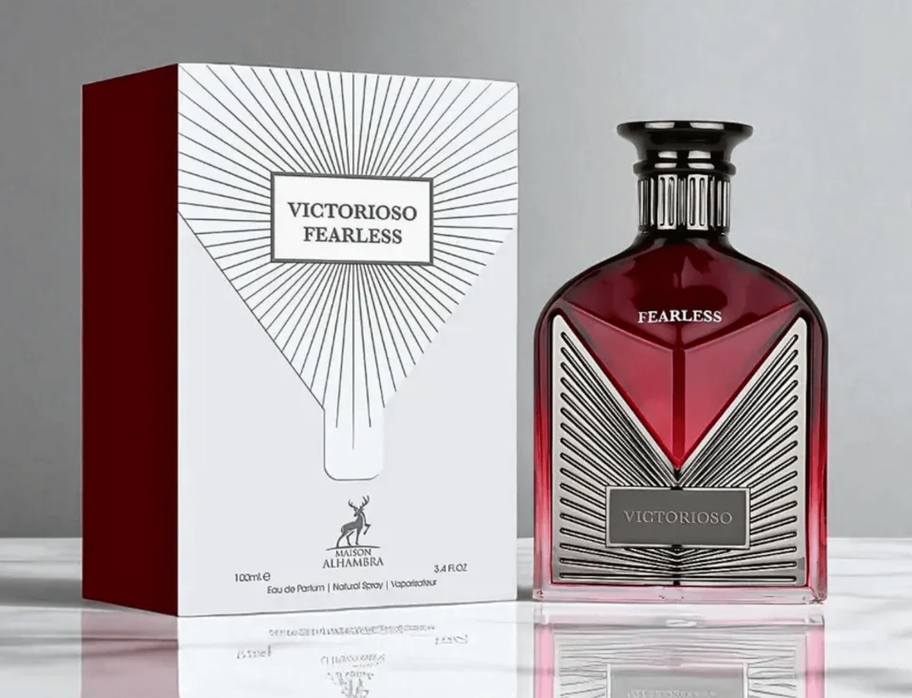 Maison Alhambra Victorioso Fearless 100ml EDP — A red glass perfume bottle with a silver geometric design and black cap, placed next to its white and red patterned box on a reflective surface.