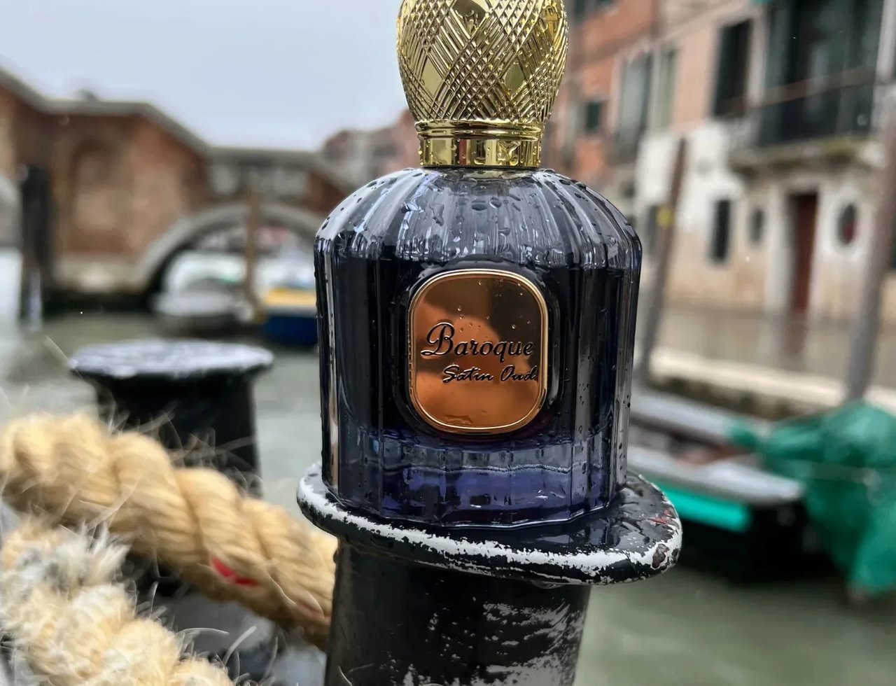 Maison Alhambra Baroque Satin Oud 100ml — Botol kaca ungu gelap dengan penutup emas bertekstur, diletakkan di atas tiang berlabuh dengan tali tebal, latar belakang terusan Venice yang kabur.