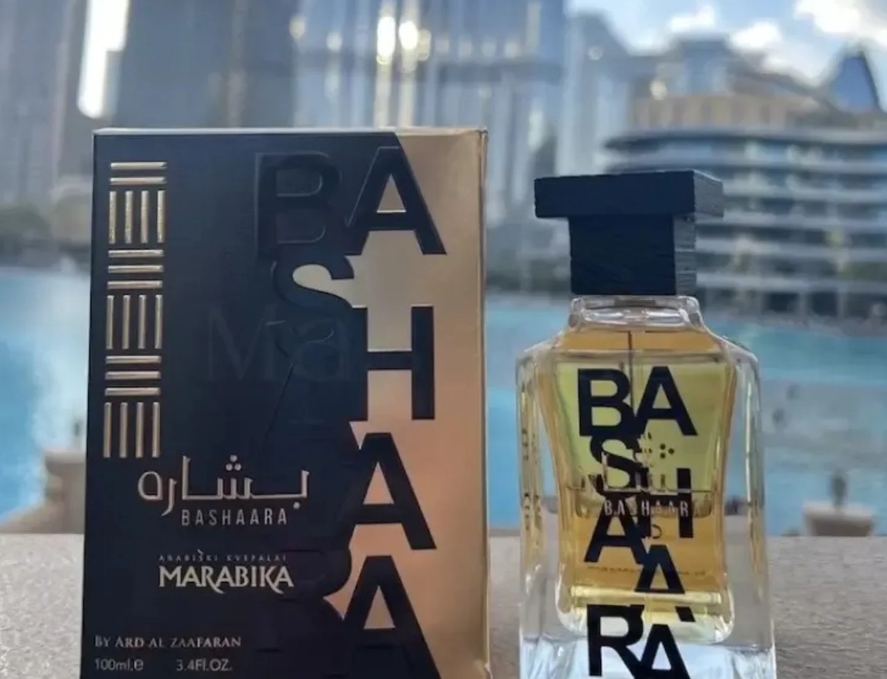 Ard Al Zaafaran Bashaara Eau De Parfum — A perfume box and bottle stand on a surface with a blurred cityscape and water in the background. | Kotak dan botol minyak wangi Ard Al Zaafaran Bashaara Eau De Parfum berdiri di atas permukaan dengan latar belakang bandar dan air yang kabur.