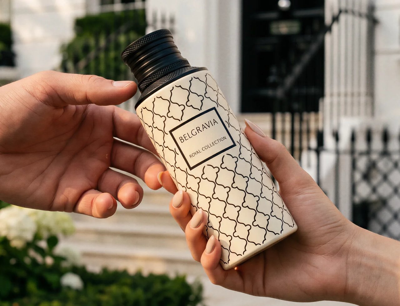 Maison Asrar Belgravia by Maison Asrar Eau de Parfum — Two hands exchanging a patterned bottle of perfume against a white building and greenery.