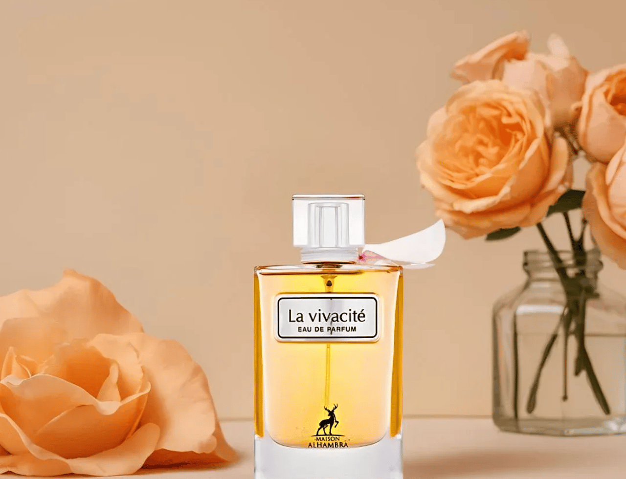 Maison Alhambra La Vivacite by Maison Alhambra 100ml EDP — Clear glass perfume bottle with golden liquid, white label, and silver cap, placed on a surface with peach roses and a vase of roses in the background.