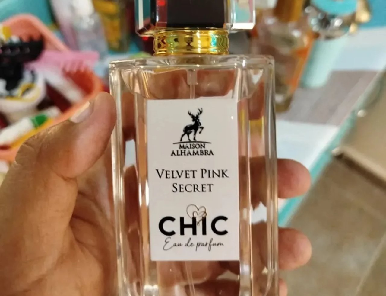 Maison Alhambra Chic Velvet Pink Secret for Women 100ml — A hand holds a clear glass perfume bottle with a white label featuring a deer logo, the text "MAISON ALHAMBRA VELVET PINK SECRET CHIC Eau de parfum" and a heart design. | Maison Alhambra Chic Velvet Pink Secret for Women 100ml — Tangan memegang botol minyak