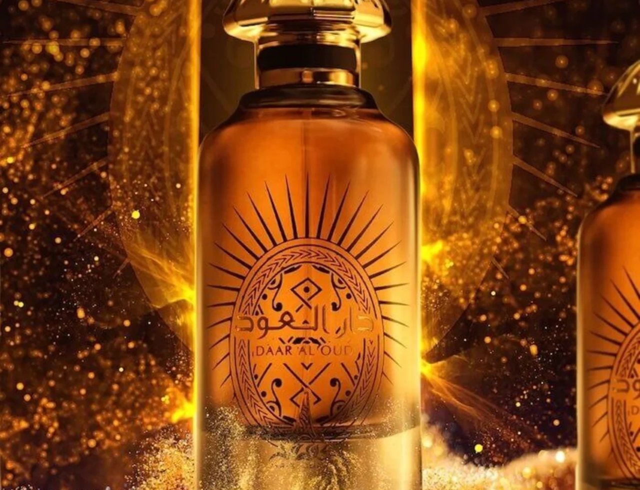 Paris Corner Daar Al Oud Intense 100ml — Botol minyak wangi berwarna keemasan dengan penutup emas berkilat, dihiasi corak matahari dan tulisan Arab. Latar belakang berkilauan dengan zarah emas.