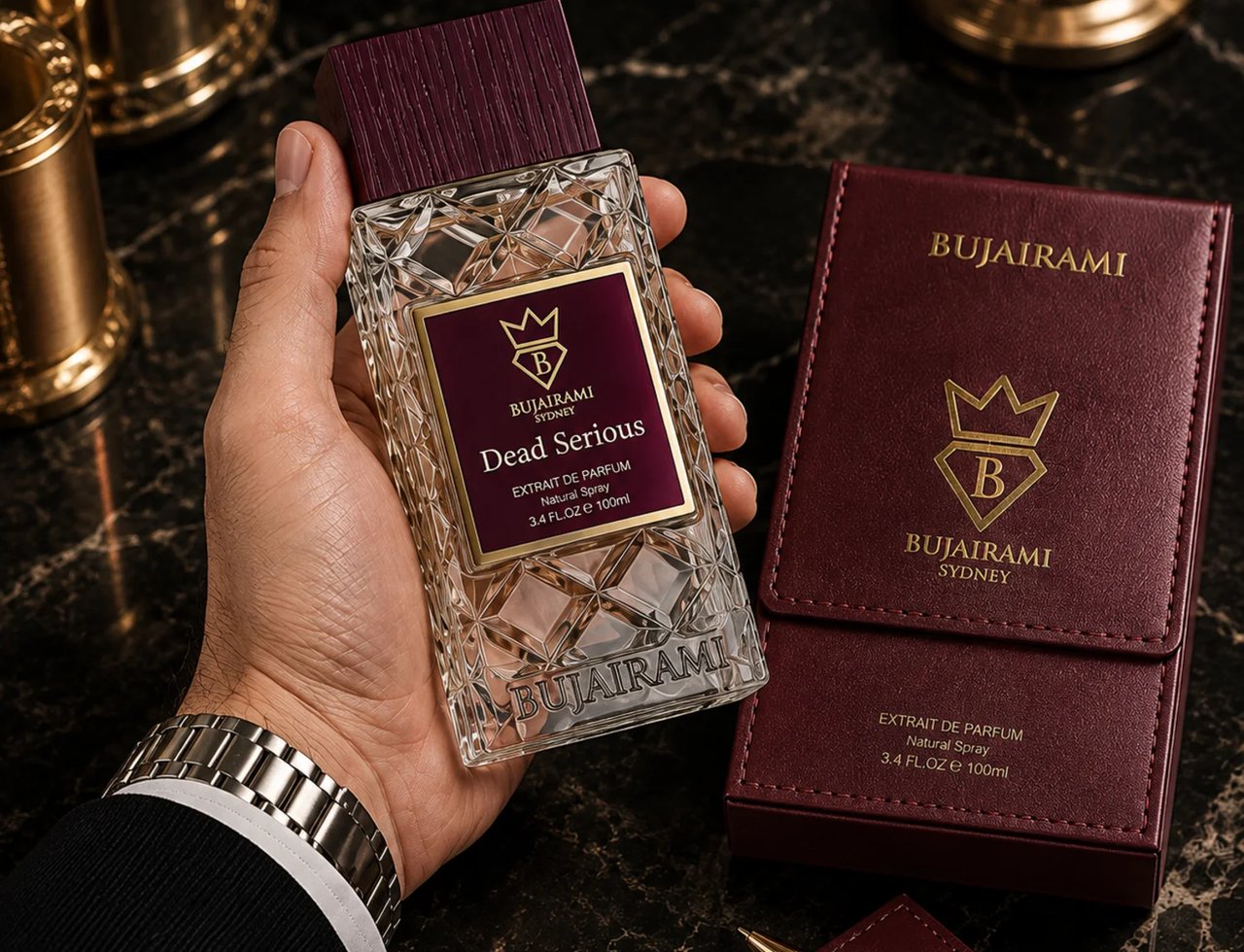 Bujairami Dead Serious by Bujairami 100ml Extrait Unisex — Hand holding a clear, faceted glass perfume bottle with a burgundy cap and label, next to a burgundy leather case and a pen on a dark marble surface.