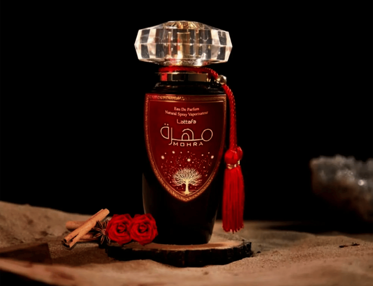 Lattafa Mohra by Lattafa 100ml EDP — Botol minyak wangi kaca gelap berlabel merah berhias reka bentuk pokok, bertutup kristal faset dan berloceng merah, diletakkan di atas hirisan kayu bersama kayu manis dan mawar merah.