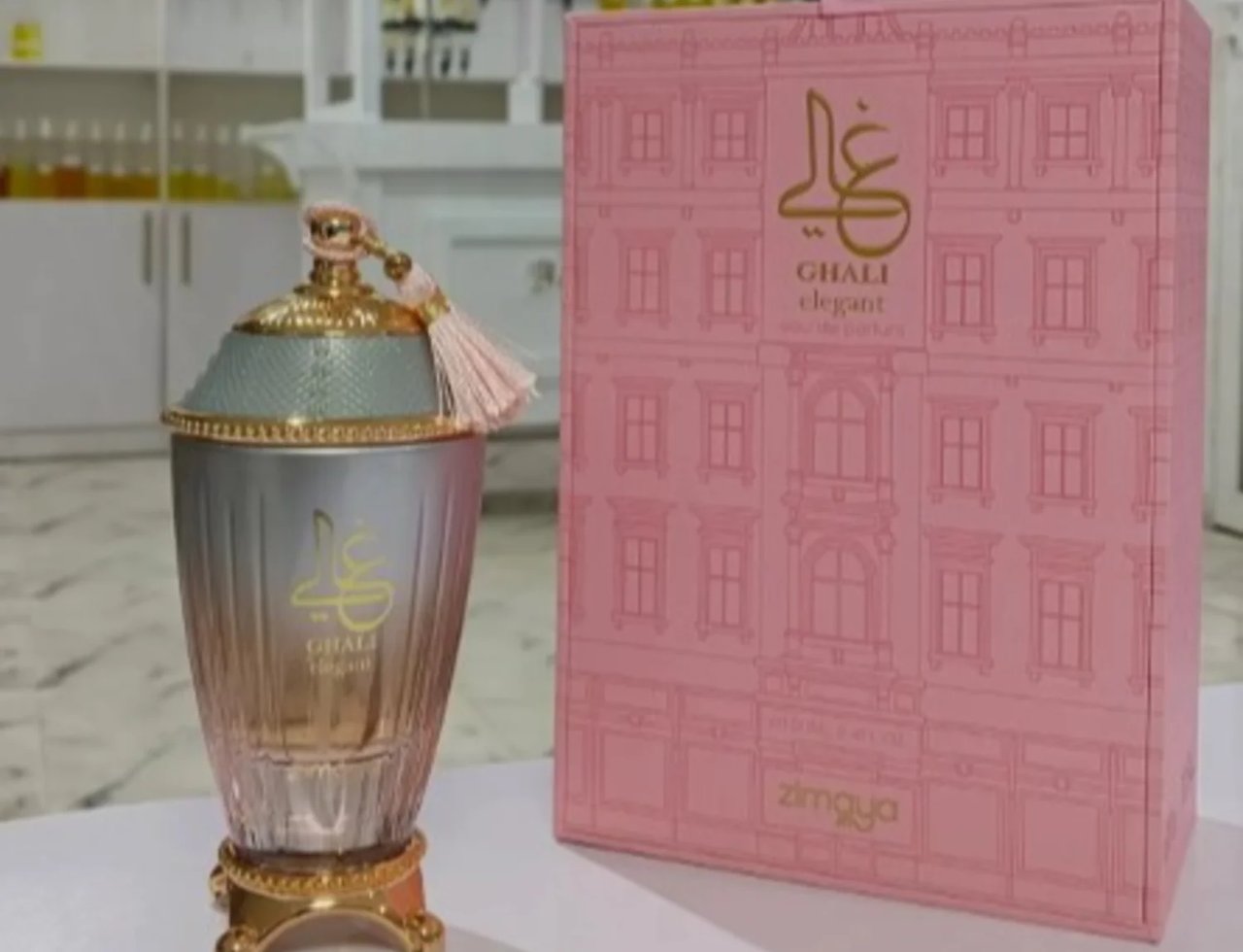 Zimaya Ghali Elegant Eau De Parfum 100ml by Zimaya — A tall, fluted glass perfume bottle with a gold base and a light blue textured cap adorned with a pink tassel, stands next to its matching pink box decorated with a building facade pattern. | Zimaya Ghali Elegant Eau De Parfum 100ml by Zimaya — Botol kaca berlekuk