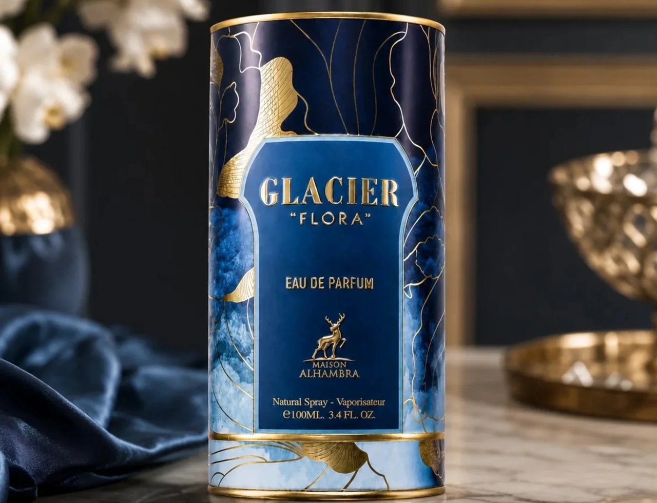 Maison Alhambra Glacier Flora 100ml — Botol silinder biru gelap berhias corak bunga emas dan rusa, diletakkan di atas meja marmar dengan kain satin biru dan pasu bunga di latar belakang.