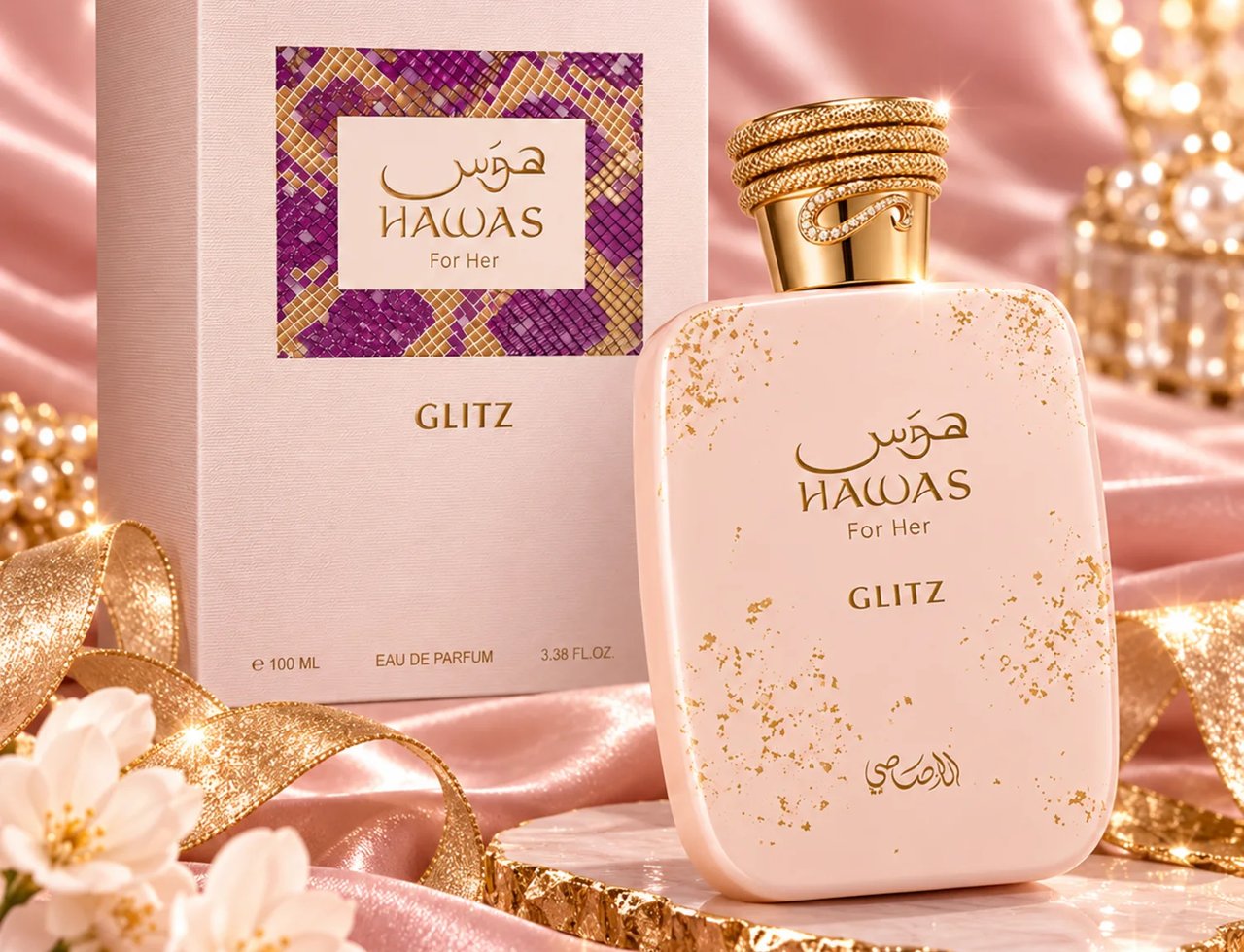 RASASI Hawas Glitz by Rasasi 100ml EDP — Botol minyak wangi merah jambu muda bertabur emas dengan penutup emas bertekstur, diletakkan di sebelah kotak merah jambu sepadan dengan sisipan corak ungu dan emas, di atas latar satin merah jambu dengan bunga putih dan reben emas.