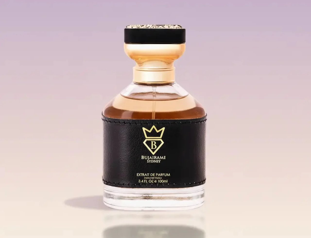 Bujairami Kingsman oleh Bujairami 100ml Extrait de Parfum — Botol minyak wangi kaca jernih bersalut seperti kulit hitam, perincian emas, dan penutup hitam berhias emas, berlatar belakang gradien lembut.