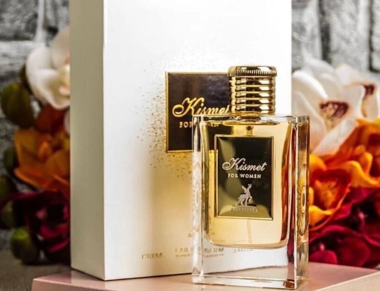 Maison Alhambra Kismet for Women 100ml by Maison Alhambra — A clear glass perfume bottle with a gold cap and gold label featuring a deer silhouette, placed next to its white and gold box, with blurred floral arrangements in the background. | Maison Alhambra Kismet for Women 100ml by Maison Alhambra — Botol minyak