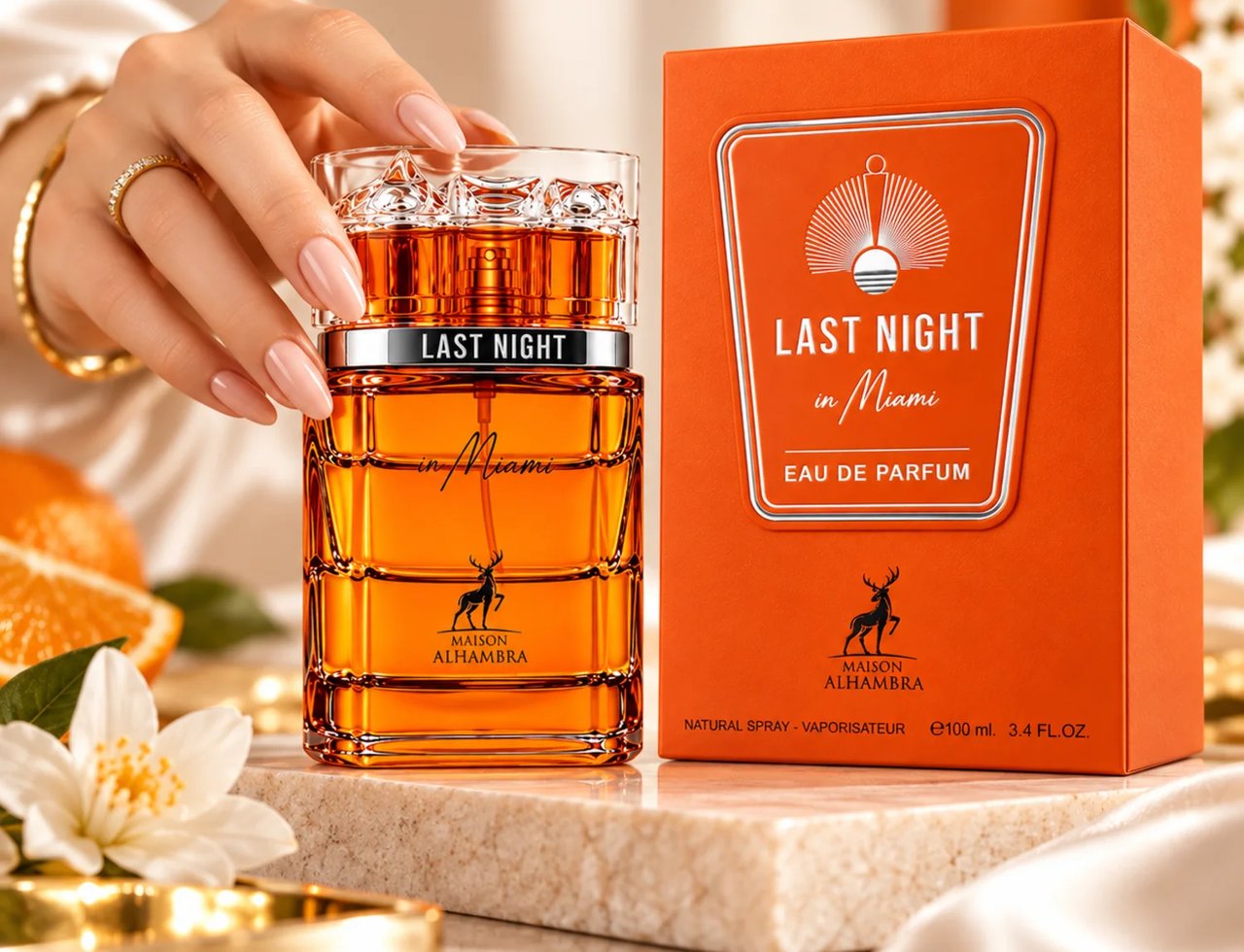 Maison Alhambra Last Night in Miami — A hand with a gold bracelet holds a clear glass perfume bottle with amber liquid. The bottle has a silver band and the box is orange.