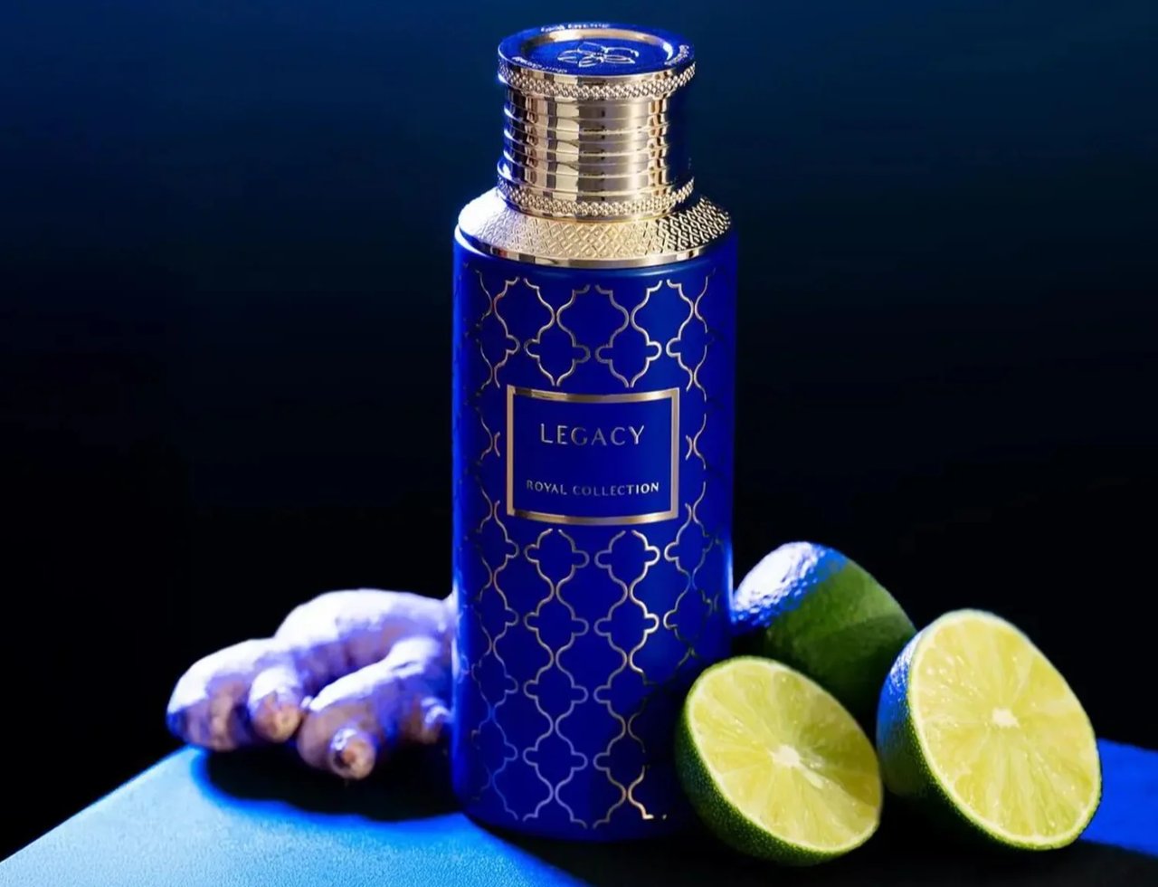Legacy by Maison Asrar 100ml EDP Unisex — Botol biru corak geometri emas, penutup emas, dan label hiasan, diletakkan di atas permukaan gelap bersama akar halia dan hirisan limau.