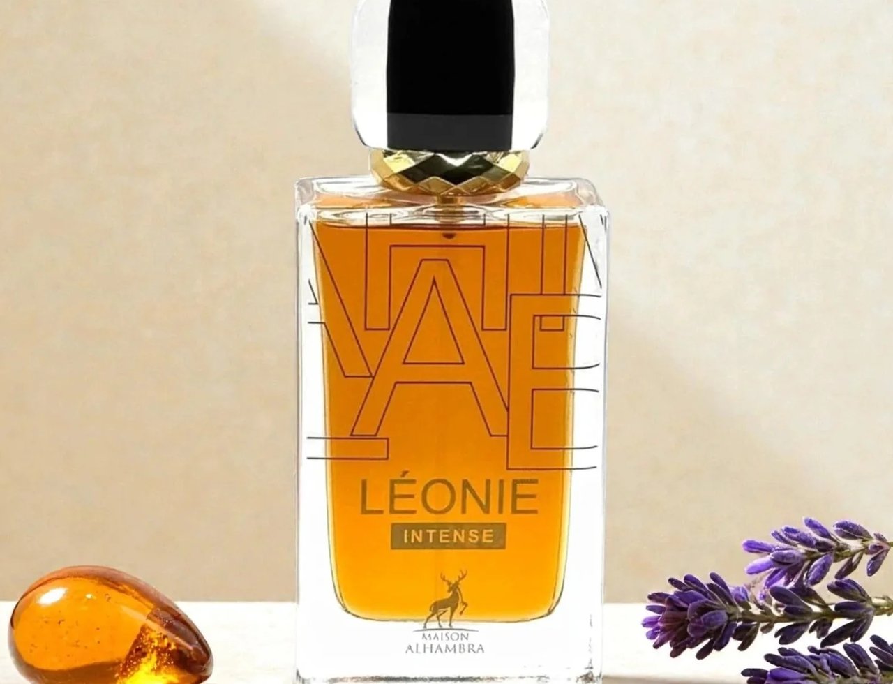 Maison Alhambra Leonie Intense — A rectangular glass perfume bottle with amber liquid, a black and gold cap, and a deer logo on the label, sits on a reflective surface next to a piece of amber and lavender sprigs. | Maison Alhambra Leonie Intense — Botol kaca berbentuk segi empat dengan cecair berwarna ambar, penutup