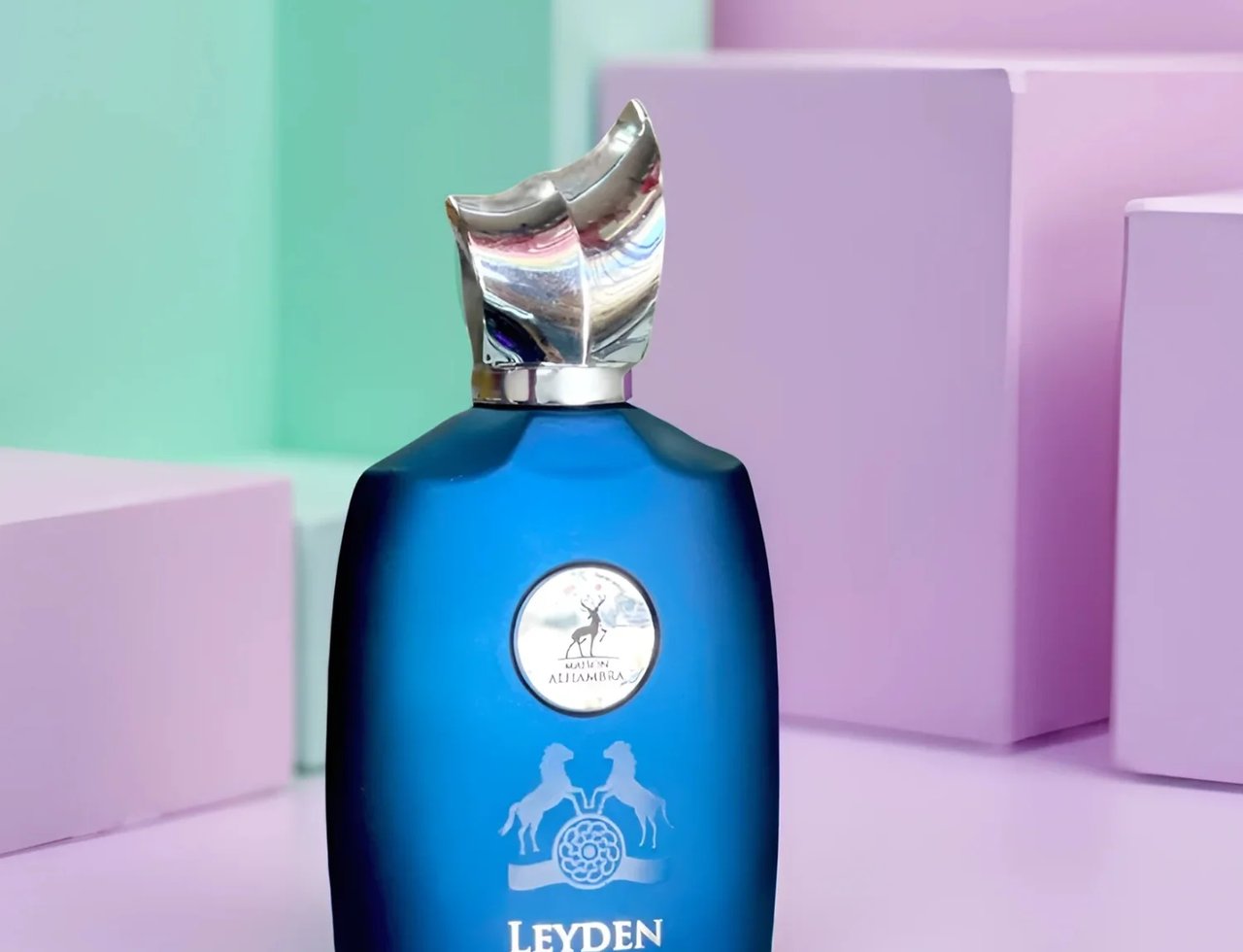Maison Alhambra Leyden by Maison Alhambra Eau De Parfum 100ml — Botol minyak wangi biru gelap matte dengan penutup perak berkilat, logo bulatan di tengah, dan tulisan 'LEYDEN' di bawah, berlatar belakang blok warna pastel ungu dan hijau.