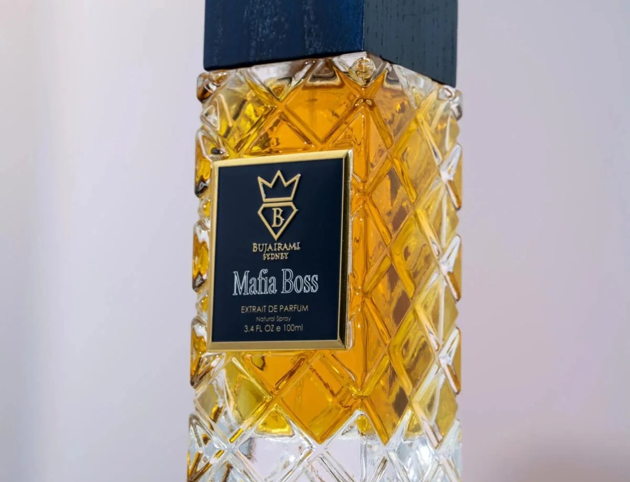 Bujairami Mafia Boss by Bujairami 100ml Extrait de Parfum — Close-up of a textured glass perfume bottle filled with golden liquid, featuring a dark blue wooden cap and a black label with gold accents.