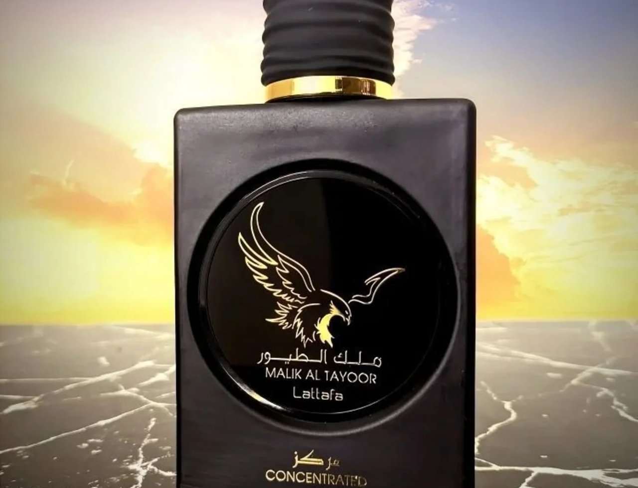 Lattafa Malik Al Tayoor Concentrated — Botol minyak wangi hitam legam dengan penutup berjalur hitam dan cincin emas, dihiasi logo helang emas di bahagian hadapan, diletakkan di atas permukaan marmar hitam berkilat dengan latar belakang langit senja berwarna jingga dan biru.