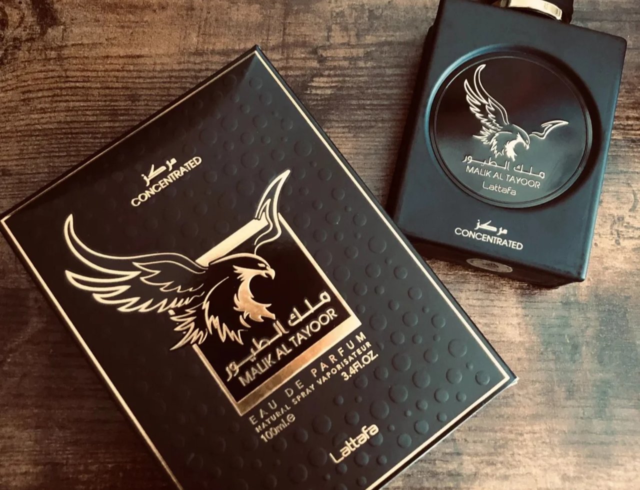 Lattafa Malik Al Tayoor Concentrated 100ml — A black perfume box with gold eagle emblem and text sits next to a black perfume bottle with a gold eagle emblem on a wooden surface. | Lattafa Malik Al Tayoor Concentrated 100ml — Kotak minyak wangi hitam dengan lambang helang emas dan teks terletak di sebelah botol minyak