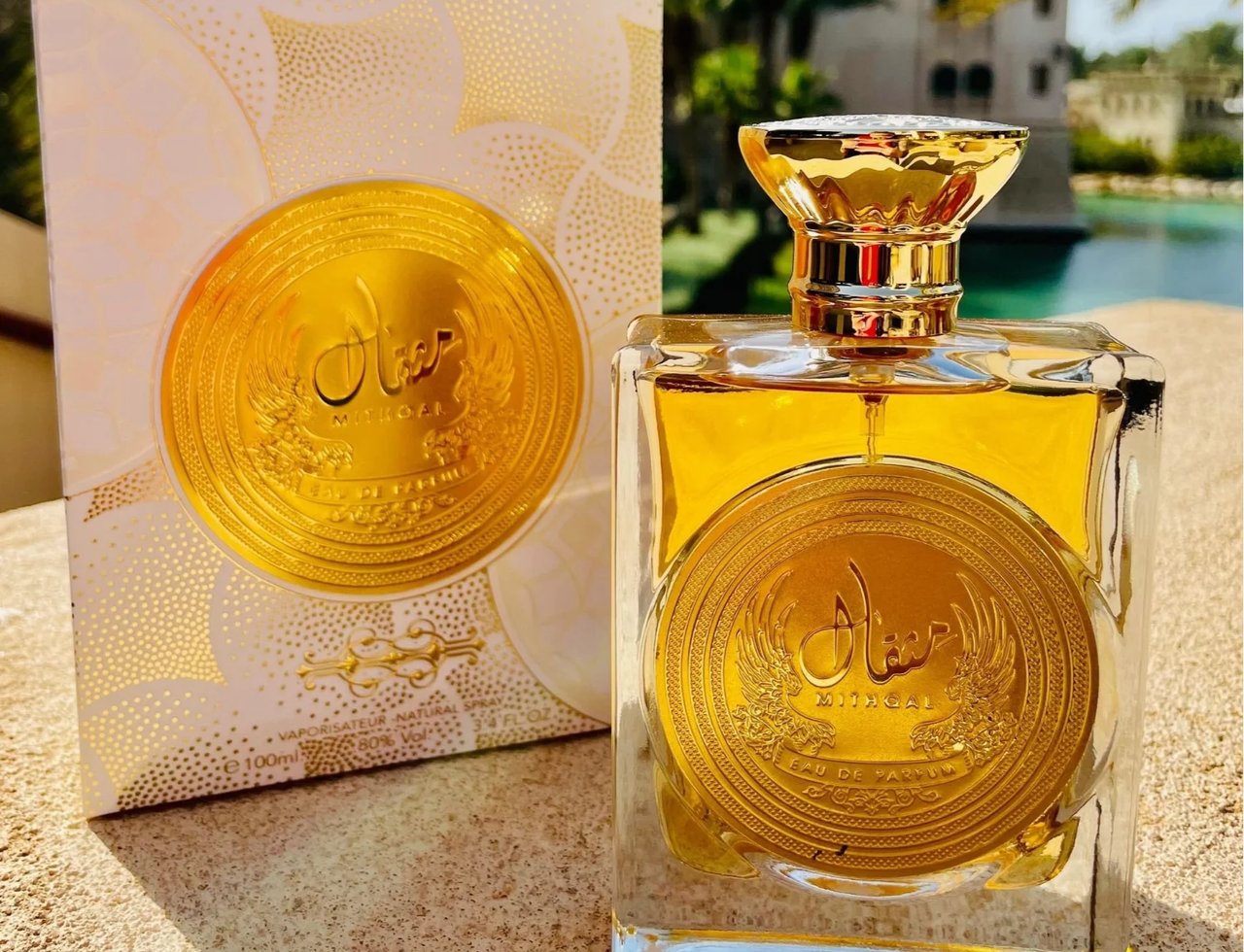 Ard Al Zaafaran Mithqal 100ml — A square glass perfume bottle with a gold emblem and golden liquid sits next to its matching white and gold patterned box, set against a blurred background of water and palm trees. | Ard Al Zaafaran Mithqal 100ml — Sebotol minyak wangi kaca berbentuk segi empat dengan lambang emas dan